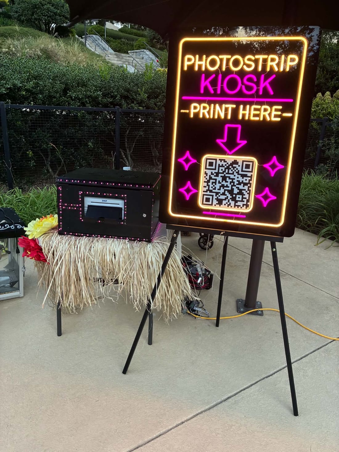 A Photostrip kiosk set up at an event: a compact printer dressed with a grass skirt next to a glowing neon sign that reads Photostrip Kiosk, Print Here, with a QR code to scan.